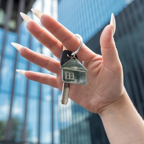 Woman hand holding new house key against the backdrop of urban development, new modern building. Real estate concept